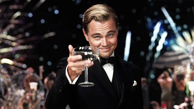 Post cover: Leonardo DiCaprio as Jay Gatsby in the movie The Great Gatsby (2013), elegantly dressed in a tuxedo, raises a glass with a confident smile against a backdrop of sparkling lights and a festive party scene.